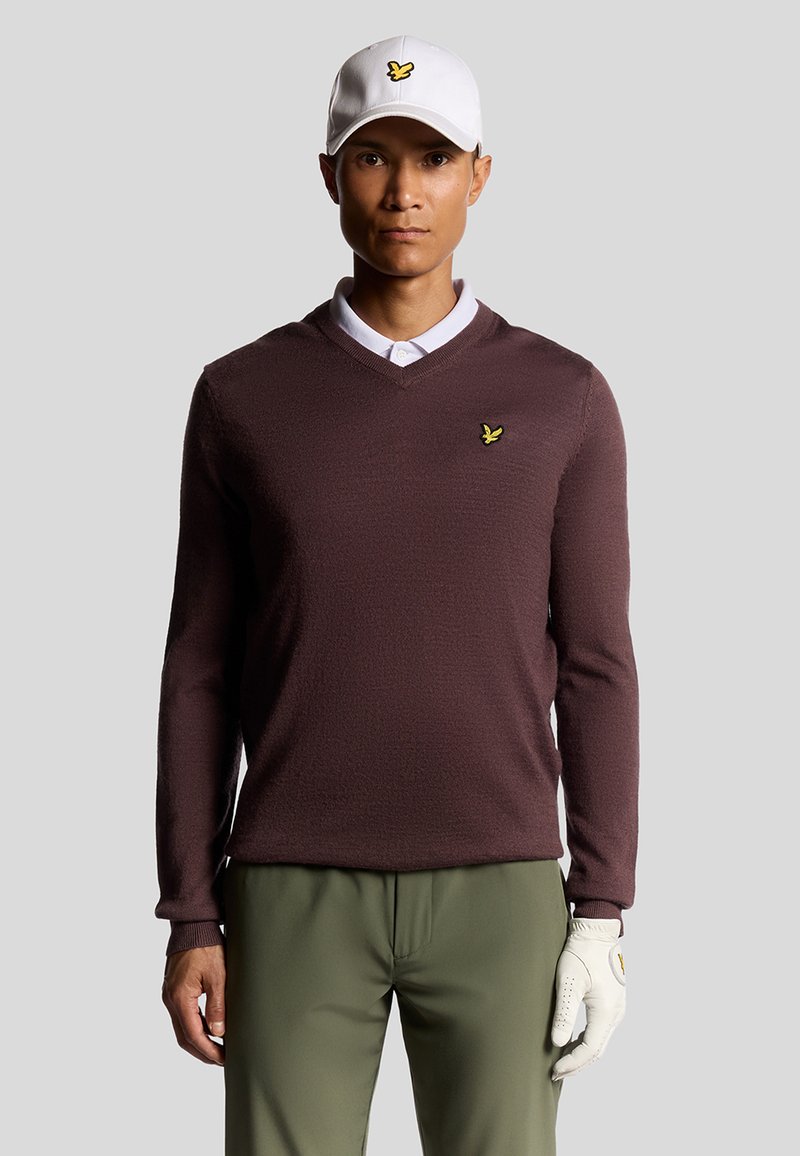 Maglione a maglia marrone con scollo a V, camicia bianca con colletto sotto, abbinato a pantaloni verde oliva e un cappellino bianco con un logo giallo.