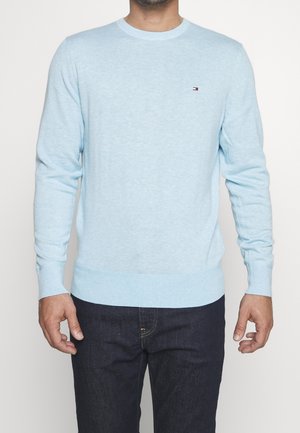 Homme portant un pull bleu clair à manches longues avec un petit logo sur la poitrine et un jean en denim foncé, debout devant un fond blanc.