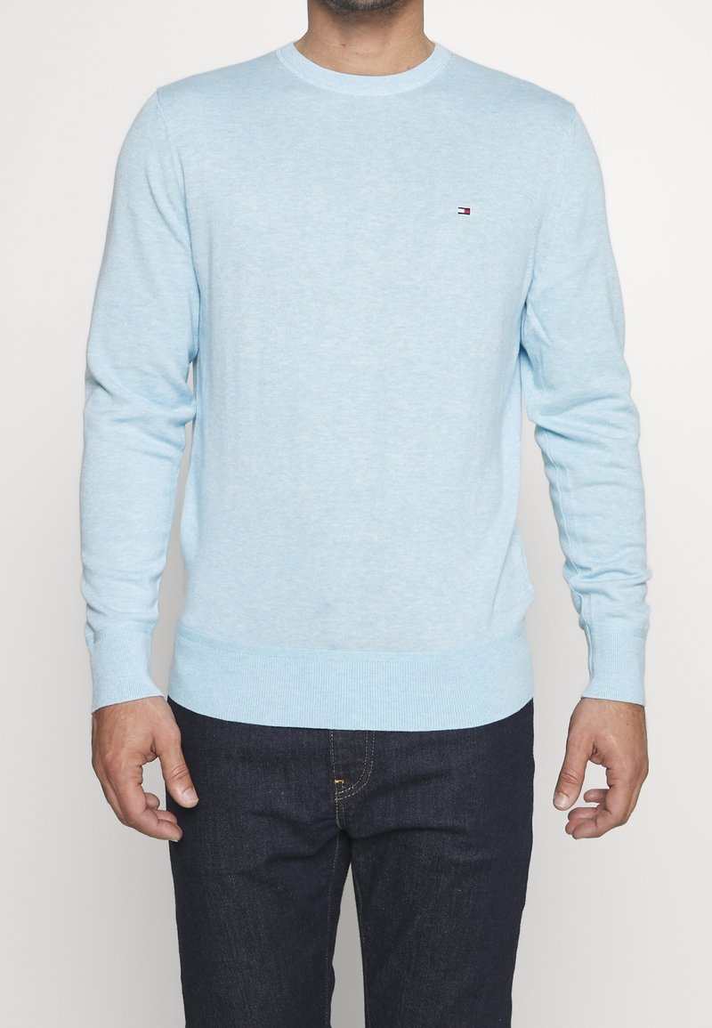 Homme portant un pull bleu clair à manches longues avec un petit logo sur la poitrine et un jean en denim foncé, debout devant un fond blanc.