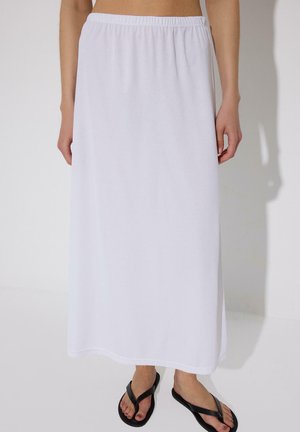 Άτομο που φορά μακριά λευκή φούστα με ελαστική μέση και μαύρα σανδάλια τύπου σαγιονάρα, όρθιο μπροστά σε απλό φόντο.