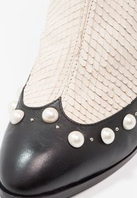 Bout avant de chaussure en cuir noir avec empeigne blanche texturée ressemblant à des écailles, décorée de gros et petits clous en perles blanches le long du bord.