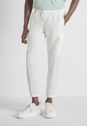 Donkerhuidig persoon draagt witte cargo joggerbroek met elastische boorden en witte sneakers, staand tegen een effen achtergrond.