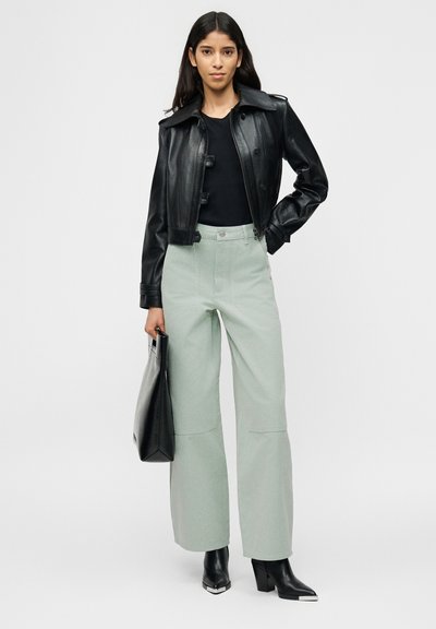 Γυναίκα που φοράει μαύρο δερμάτινο μπουφάν, μαύρο τοπ, ανοιχτό γκρι παντελόνια με φαρδιά μπατζάκια, μαύρες μυτερές μπότες, κρατώντας μαύρη τσάντα tote, στέκεται μπροστά σε απλό φόντο.