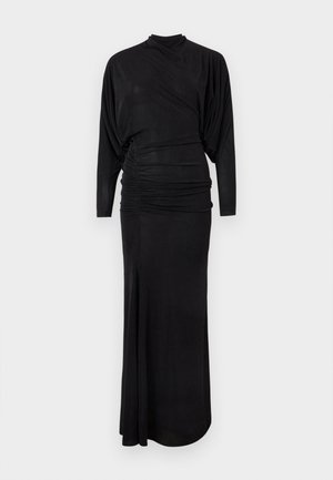 Ženska stoji z rokami na bokih, oblečena v dolgo črno obleko s krili, podobnimi netopirju, in črne odprte pete na enobarvni beli podlagi.