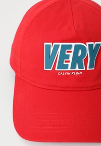 Rode katoenen pet met een gestructureerd ontwerp, voorzien van een grote blauwe "VERY" grafiek en het "CALVIN KLEIN" logo in het wit. Gladde textuur.