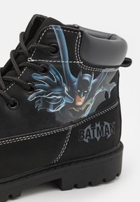 Černé semišové boty se znakem Batmana, bílým šitím, kulatými tkaničkami a texturovanou gumovou podrážkou s černým logem.