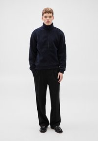 Meresinine fliis-topp, millel on kõrge krae, täispikk eesmine lukk ja soonilised varrukaäärised, millele on lisatud mustad laiade säärtega püksid ja mustad jalanõud.