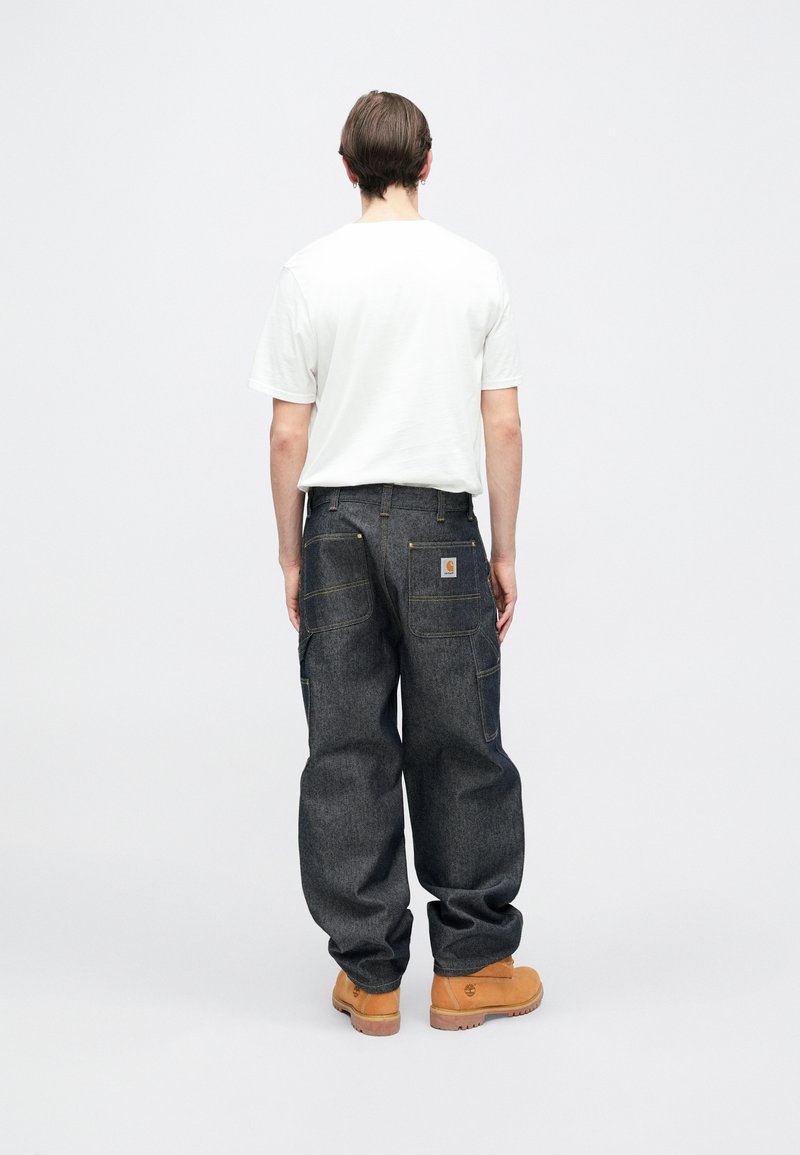 Mørk denim bukser med en løs pasform, der har to baglommer og gule syningsdetaljer. Båret med en hvid t-shirt og beige støvler.