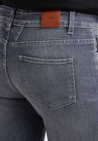 Vaqueros de mezclilla gris oscuro con una superficie texturizada, que cuentan con un bolsillo trasero y una etiqueta de marca de cuero marrón con la inscripción "CLOSED" en la cinturilla.