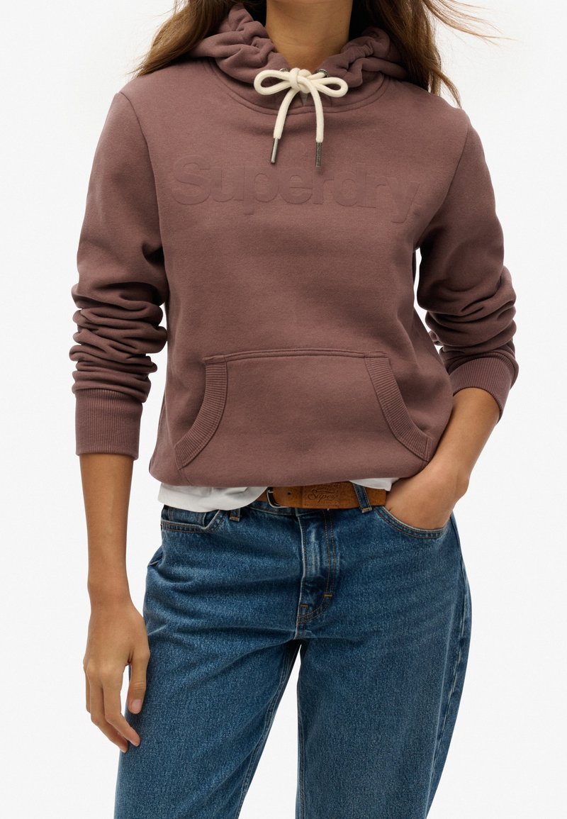 Personne portant un sweat à capuche mauve Superdry avec des cordons blancs, un jean bleu, et une main dans la poche, visible du cou aux hanches.