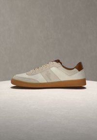 Baskets beige et blanches à profil bas avec fermeture à lacets, accents marron au col de la cheville, et semelle en gomme sur fond neutre.