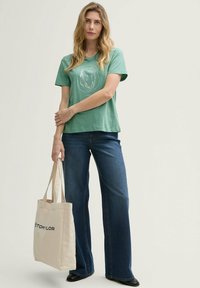 T-shirt en coton vert avec un design graphique circulaire blanc, associé à un jean évasé bleu. Sac fourre-tout beige avec logo, chaussures noires.