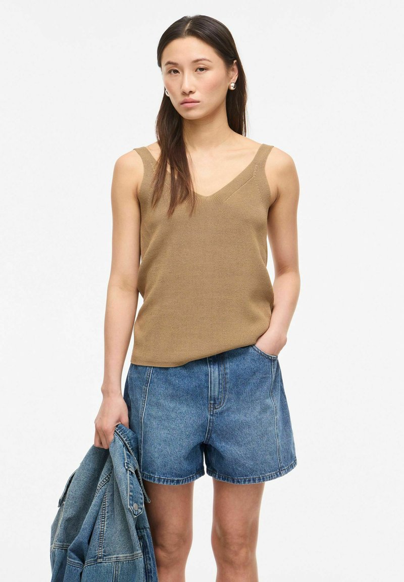 Femme portant un haut sans manches en maille beige et un short en denim, tenant une veste en denim dans sa main gauche.