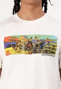 Balta medvilnės marškinėliai su spalvinga grafika, vaizduojančia kaubojus ir rančų scenas. Virš dizaino pateikta tekstas "LEVI'S".