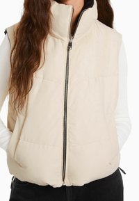 Beige puffer vest with high collar, front zipper, and side pockets. Soft, textured fabric with subtle stitching detail. Black inner lining.