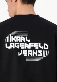 Sweatshirt preto com texto branco a dizer "KARL LAGERFELD JEANS." Apresenta um design gráfico moderno, incluindo riscas e variação na espessura das letras.