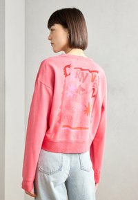 Sudadera corta rosa con un diseño gráfico en la parte posterior que presenta formas y patrones audaces. Puños y dobladillo acanalados. Tejido ligero.