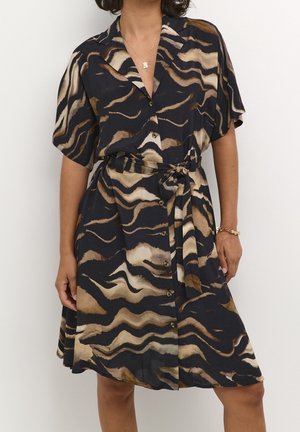 Femme portant une robe à boutons devant, à manches courtes, avec un motif abstrait en noir, beige et marron, et une ceinture nouée à la taille.