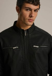 Czarna skórzana kurtka z wysokim kołnierzem, wyposażona w dwie zamykane na zamek kieszenie na piersi oraz gładką teksturę. Delikatny połysk podkreśla design.