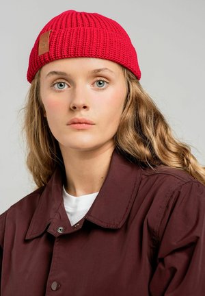 Czerwona czapka z dzianiny o prążkowanej strukturze i skórzanej metce. Noszona z marsala koszulową kurtką, na białej bluzce. Neutralne tło.