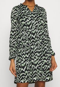 Langärmeliges Kleid in Schwarz und Grün, mit einem Zick-Zack-Muster, V-Ausschnitt und gefaltetem Oberteil, aus leichtem, strukturiertem Stoff.