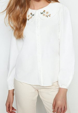 Vrouw draagt een witte blouse met knopen en een geborduurde ronde kraag met bloemenmotief en lange mouwen, gecombineerd met lichtbeige broek.