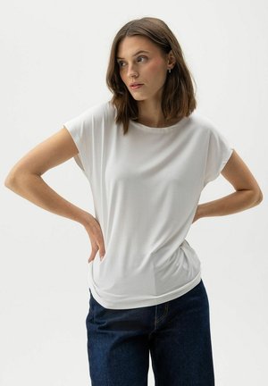 Noorpruunide õlgadeni juustega noor naine, kes kannab valget lühikeste varrukatega tuba ja tume siniseid teksapükse, käed puusadel, vaadates kõrvale.