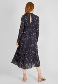 Lange marineblauwe jurk met bloemenpatroon, geplooide rok, sleutelgatdetail op de rug en lange doorschijnende mouwen. Gecombineerd met lichtgekleurde blokhak sandalen.
