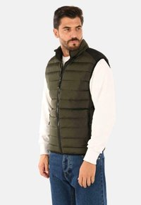 Gilet verde imbottito con colletto alto, chiusura con zip e dettagli neri. Due tasche laterali e orlo dritto. Indossato sopra una camicia bianca.