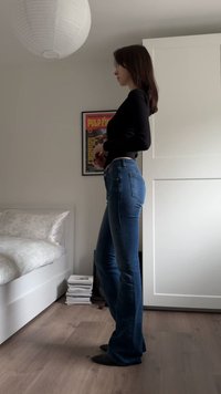 Jean flare bleu avec une finition délavée, porté avec un haut noir à manches longues. Tissu lisse, coupe ajustée, et légère élasticité. Fond en parquet bois.