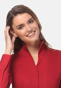 Blouse rouge à col avec des manches longues, texture lisse et poignets boutonnés. Le col est droit, mettant en avant un design minimaliste et une couleur vive.