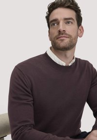 Bordeauxfarbener Rundhalsausschnitt-Pullover, gerippter Kragen und Bündchen, gestrickte Textur, getragen über einem weißen Hemd mit Kragen, vor einem neutralen Hintergrund.