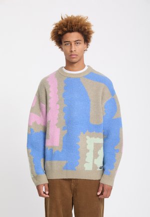 Jeune homme aux cheveux bouclés portant un pull beige avec des motifs abstraits bleus, roses et vert clair et un pantalon en velours côtelé marron, debout sur fond blanc.