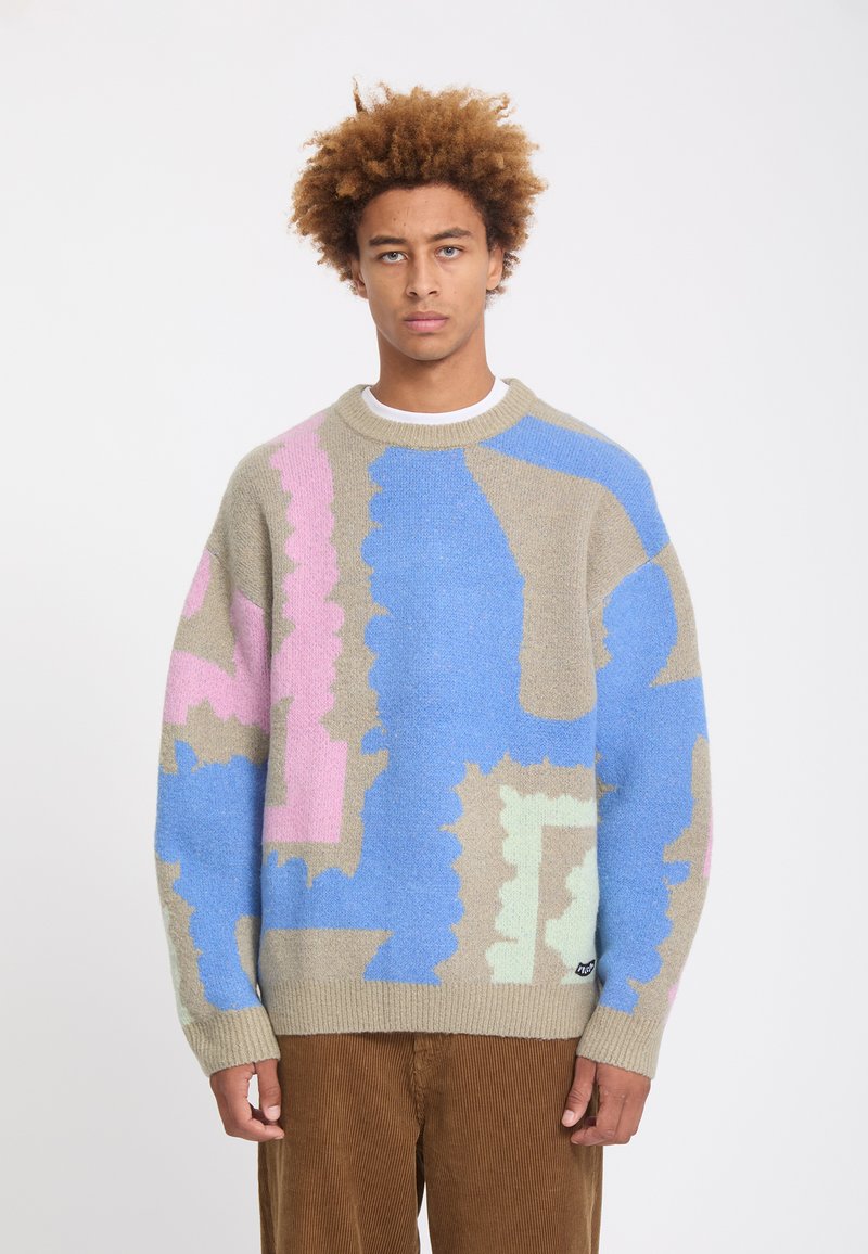 Jeune homme aux cheveux bouclés portant un pull beige avec des motifs abstraits bleus, roses et vert clair et un pantalon en velours côtelé marron, debout sur fond blanc.