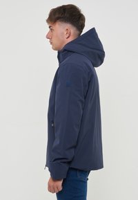 Veste étanche bleu marine avec capuche, fermeture éclair et poches latérales. Le matériau semble lisse avec une finition mate.