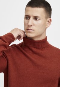 Pulover cu guler înalt, de culoare ruginită, cu textură cu nervuri, cu o siluetă ajustată. Materialul pare moale și tricotat.