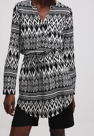 Femme portant une tunique à manches longues à motifs noir et blanc avec une taille à cordon et un short noir.
