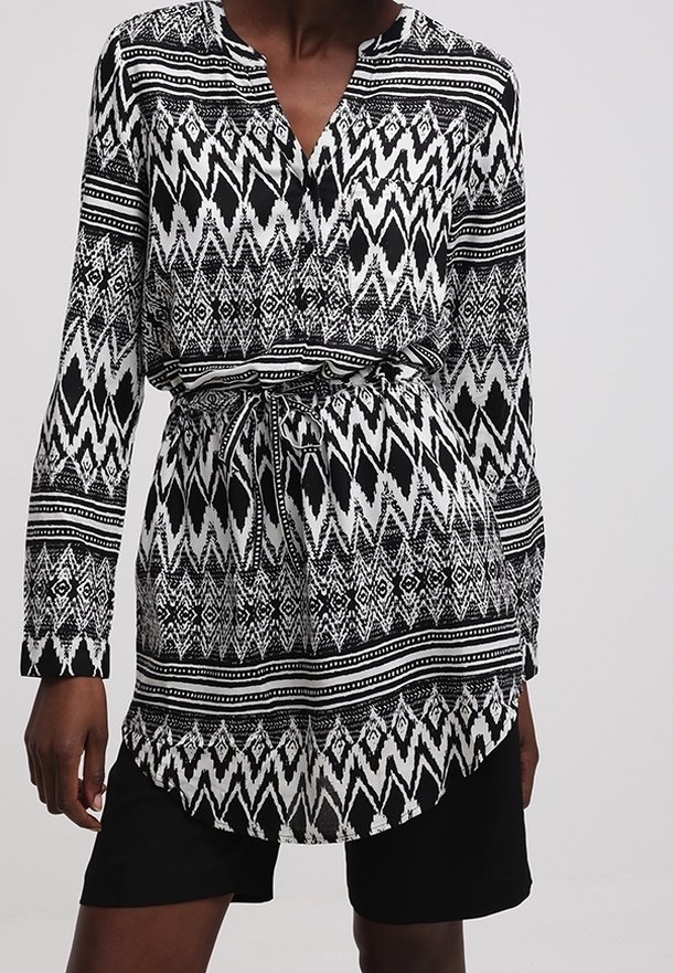 Femme portant une tunique à manches longues à motifs noir et blanc avec une taille à cordon et un short noir.