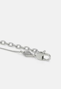 Silverkedja armband med ovala länkar, med en kräftklor och en hexagonal tagg märkt "Fossil." Slät metalltextur och polerad finish.