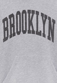 Tissu gris avec de grandes lettres majuscules noires formant BROOKLYN en arc au-dessus d'une poche kangourou.