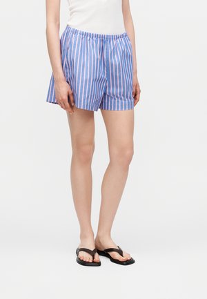 Γυναίκα που φορά φαρδιά μπλε και ροζ κάθετα ριγέ σορτς, λευκό τοπ και μαύρα σανδάλια flip-flop σε μονόχρωμο φόντο.