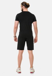 Schwarzes Baumwoll-T-Shirt mit kurzen Ärmeln, kombiniert mit schwarzen Shorts. Der Mann steht mit dem Rücken zur Kamera und trägt weiße Sneakers mit roten Akzenten.