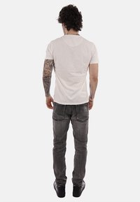 Homme aux cheveux bouclés et foncés, avec des tatouages sur le bras droit, portant un t-shirt blanc, un jean noir délavé et des baskets noires, vu de dos.