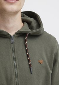 Sweat à capuche vert avec fermeture éclaire à l'avant, tissu texturé, un cordon de serrage en tresse contrastante et un petit patch logo en cuir triangulaire.