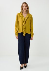 Blusa gialla senape con dettagli a volant sul davanti e maniche lunghe, abbinata a jeans a gamba larga blu scuro e tacchi a punta neri.