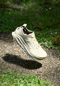Chaussure de sport beige clair avec une tige texturée, des perforations et une semelle épaisse. Présente un tirant et un logo visible sur le côté.