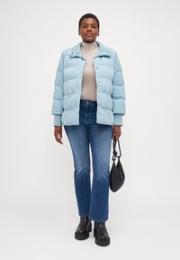 Chaqueta acolchada de color azul claro con un diseño ligeramente cropped y cierre de botones en la parte frontal, combinada con un suéter de cuello alto gris y jeans oscuros de corte acampanado.