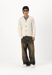 Cardigan en maille torsadée crème avec un col en V, des boutons en bois et une poche frontale, associé à un jean bleu délavé ample et des baskets blanches.