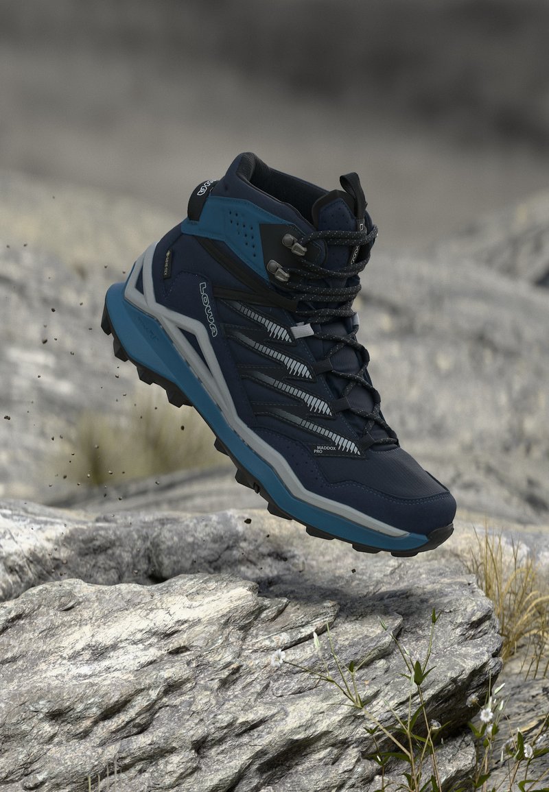 Laivastonsininen ja harmaa vaelluskenkä, jossa on jyrkkä pohja, ilmassa kivenlaisen maaston yllä, jossa on niukasti ruohoa ja pieniä valkoisia kukkia.