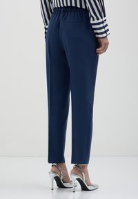Marineblaue Hosen mit einem breiten elastischen Bund, tailliert geschnitten und zwei Gesäßtaschen, kombiniert mit silbernen High-Heels.